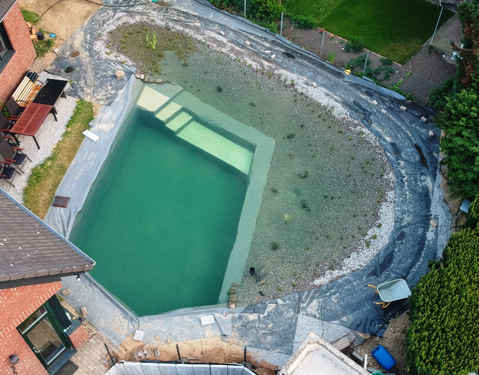 Luftbild zeigt den fortschreitenden Bau des Schwimmteichs bei Mielke's Naturbadeteich. Die Teichabdichtung ist abgeschlossen, Treppe und Plateau mit Anti-Rutsch-Folie sind sichtbar. Das Wasser hat einen hohen Pegel erreicht, und Wasserpflanzen wurden gepflanzt.