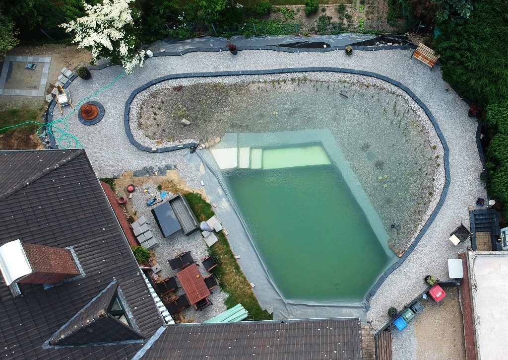 Luftbild zeigt den fortschreitenden Bau des Schwimmteichs bei Mielke's Naturbadeteich. Die Teichabdichtung ist abgeschlossen, Treppe und Plateau mit Anti-Rutsch-Folie sind sichtbar. Die Randgestaltung mit Trockenmauer dient zusammen mit der Folie als Kapillarsperre. Rings um den Teich wurde Kies als Weg angefüllt, der für perfekte Randgestaltung und Wege sorgt.