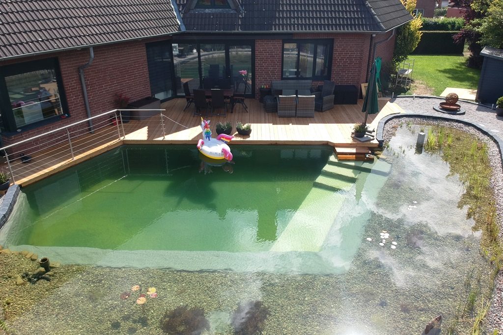 Bild zeigt den fertigen Schwimmteich bei Mielke's Naturbadeteich, wo ein aufblasbares Einhorn für Spaß sorgt. Die Terrasse ist komplett. Erste Pflanzen sprießen.
