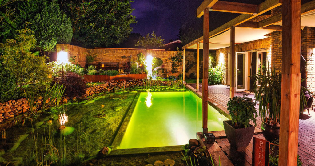DIY Schwimmteich bei Nacht mit Beleuchtung, gewachsene Pflanzen, klares Wasser, und eine Pergola über der Terrasse.
