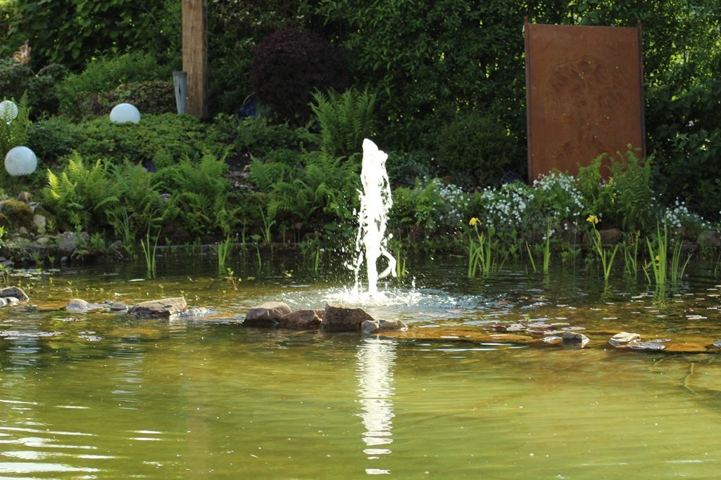 Eine Wasserfontäne schießt Wasser aus dem Pflanzenbereich in die Luft und sorgt so für ein spannendes Ambiente. Im Hintergrund sind rostige Dekoelemente aus Metall