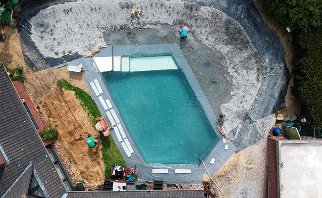 Luftbild zeigt einen im Bau befindlichen Schwimmteich von Mielke's Naturbadeteich. Das Wasser erreicht den Regenerationsbereich und bedeckt teilweise den Kies. Es werden Wasserpflanzen gepflanzt.