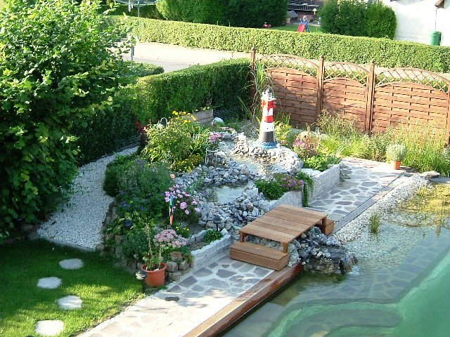 Liebevoll dekorierter Bachlauf neben einem Schwimmteich gebaut aus vielen kleinen Natursteinpflanzen Leuchtturm und einer Treppe