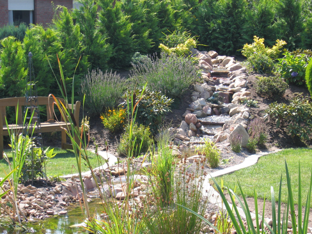 Bachlauf abgehend von einem Hügel gestaltet mit mehreren Natursteinen er mündet in den Pflanzenbereich