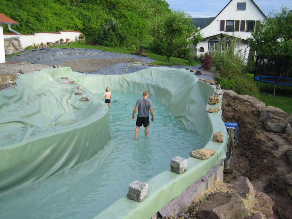 Der Schwimmbereich eines individuellen Schwimmteichs, der mit grüner Folie von Mielke's Naturbadeteich ausgekleidet wurde, wird mit Wasser befüllt. Im Hintergrund ist die Hanglage und ein Haus zu sehen.