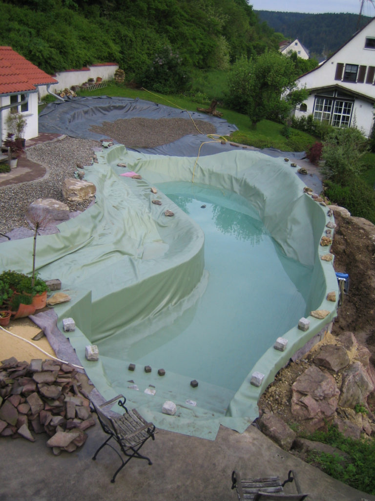 Eine Treppe mit grüner Folie im Schwimmbereich eines individuellen Schwimmteichs, der mit Folie von Mielke's Naturbadeteich gestaltet wurde. Die Stufenkanten sind provisorisch mit Steinen beschwert. Eine Steinterrasse und ein Kiesbett sind ebenfalls sichtbar.