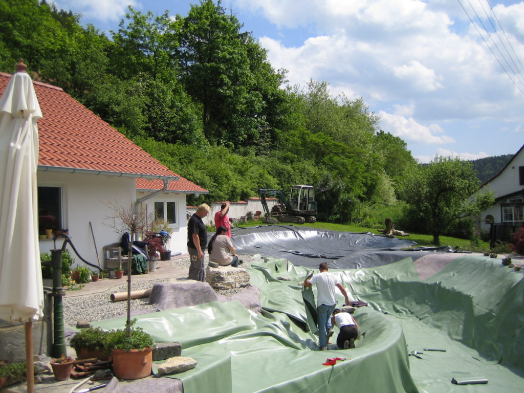 Die linke Pflanzzone eines individuellen Schwimmteichs, der mit grüner Folie von Mielke's Naturbadeteich ausgekleidet wurde. Auf der linken Seite befindet sich ein Kiesbett und ein Haus