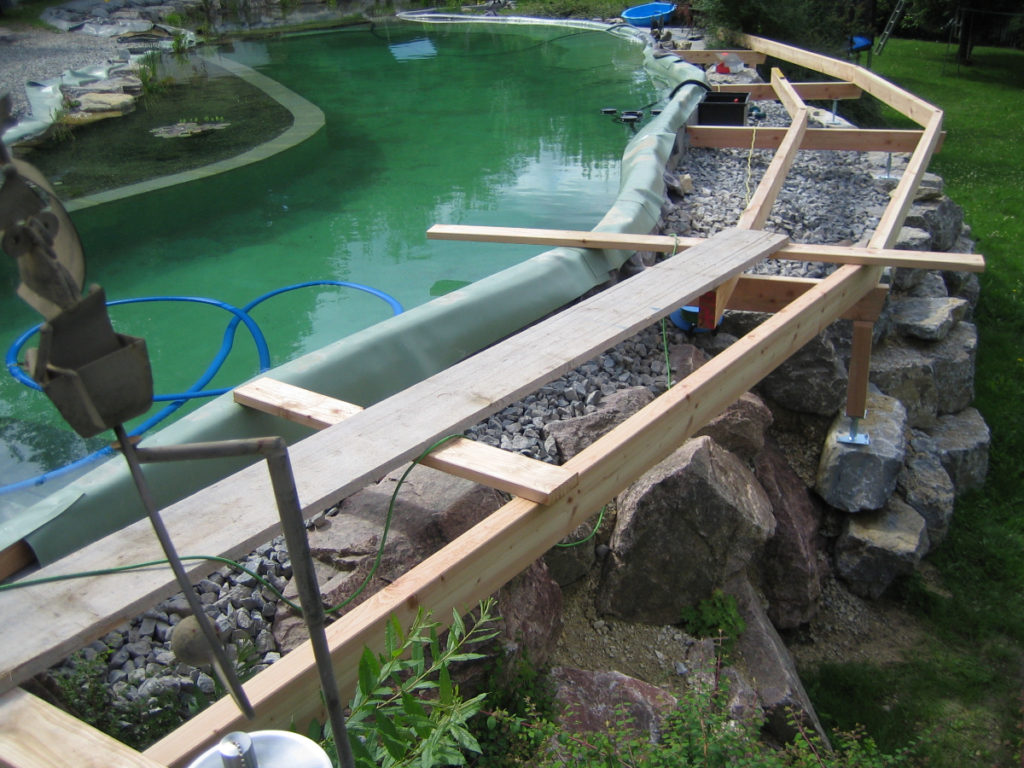 Eine Unterkonstruktion aus Holz, abgestützt auf Felssteine, umgibt den Schwimmbereich eines individuellen Schwimmteichs, der mit Folie von Mielke's Naturbadeteich selber gebaut wurde.