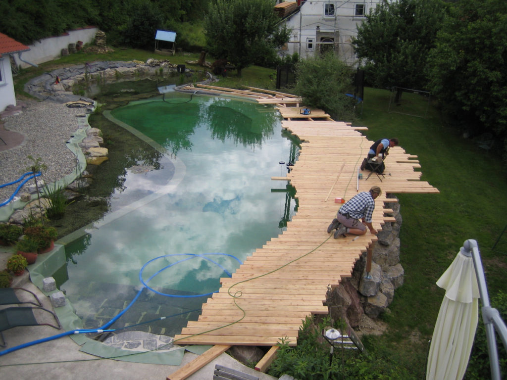 Zwei Personen verschrauben Bretter auf einer Unterkonstruktion im Schwimmbereich eines individuellen Schwimmteichs. Die Treppe wurde mit Steinen belegt, und Pflanzen im Kiesbett sind zu sehen.