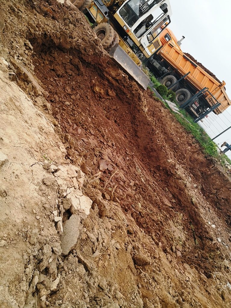 Ein Bagger baggert Erde aus, während im Hintergrund ein LKW mit abgetragener Erde steht.