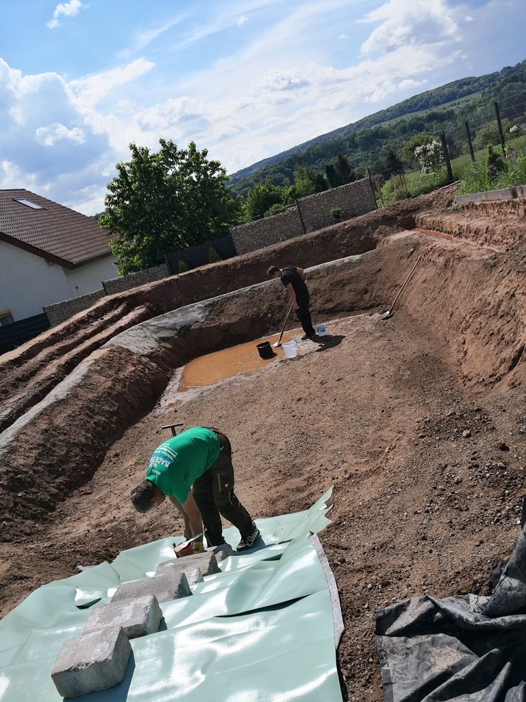 Eine Mitarbeiterin entfernt Regenwasser vom Schwimmbereich, während ein Mitarbeiter von Mielke's Naturbadeteich grüne Folie auf den Treppenstufen auslegt und mit Steinen beschwert.