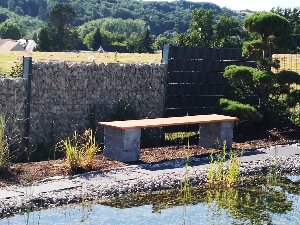 Eine Bank aus Stein und Holzbrett steht am Wegrand mit Blick auf den Design-Schwimmteich mit seinem außergewönlichem Ausblick