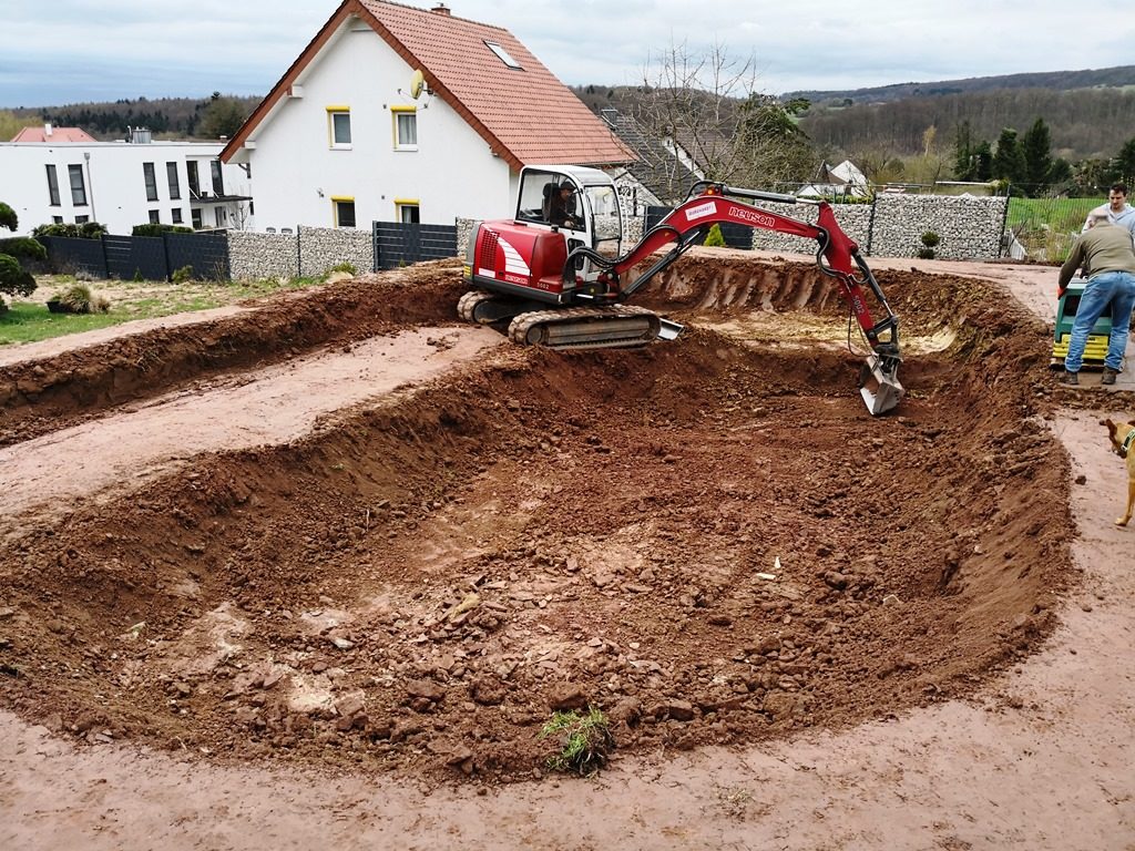 Ein Bagger formt einen ovalen Erdaushub mit einem Plateau, während zwei Arbeiter die Erde am Rand verdichten.