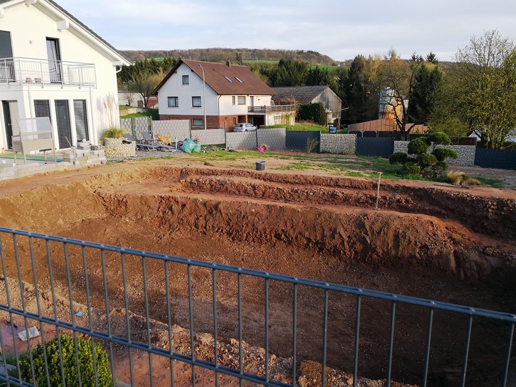 Seitenansicht des ausgehobenen Schwimmteichs mit einer ovalen Form und abgestuften Seiten, im Hintergrund sind Häuser zu sehen.