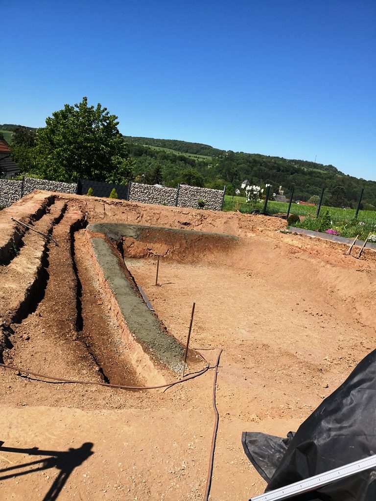 Vorderansicht des ausgehobenen Schwimmteichs, mit Metallstangen zur Abgrenzung des Schwimmbereichs und einer treppenförmigen Pflanzzone in U-Form. Ein außergewöhnlich schöner Ausblick auf die Landschaft.