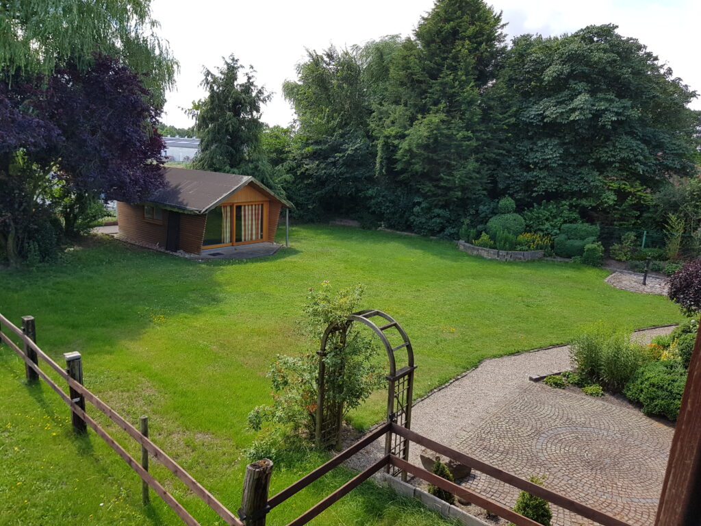 Rasenfläche mit Holzhütte, Zaun, Steinweg und Torbogen