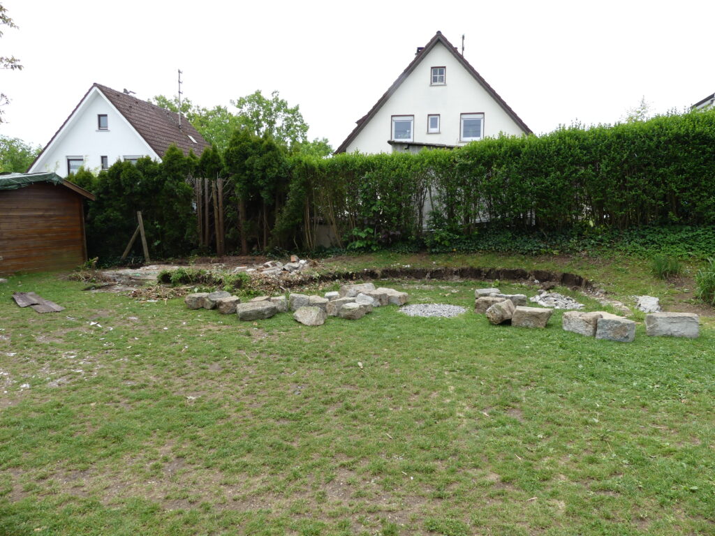 Gestaltung des Gartenbereichs um Mielke's Naturbadeteich: Steine, Holzhütte und Hecke auf einer grünen Wiese.