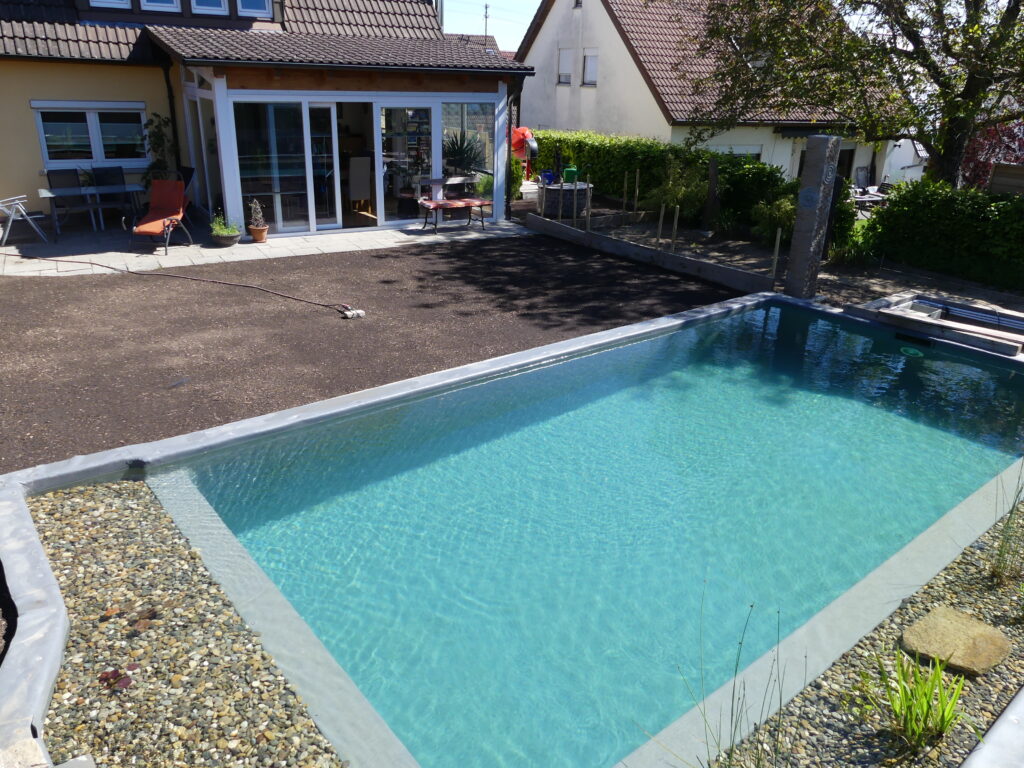 Der Naturpool wurde komplett mit Wasser befüllt, in der Filterzone stehen einige Wasserpflanzen, eine freie Fläche befindet sich neben dem Naturpool, dahinter eine Steinterasse mit Haus