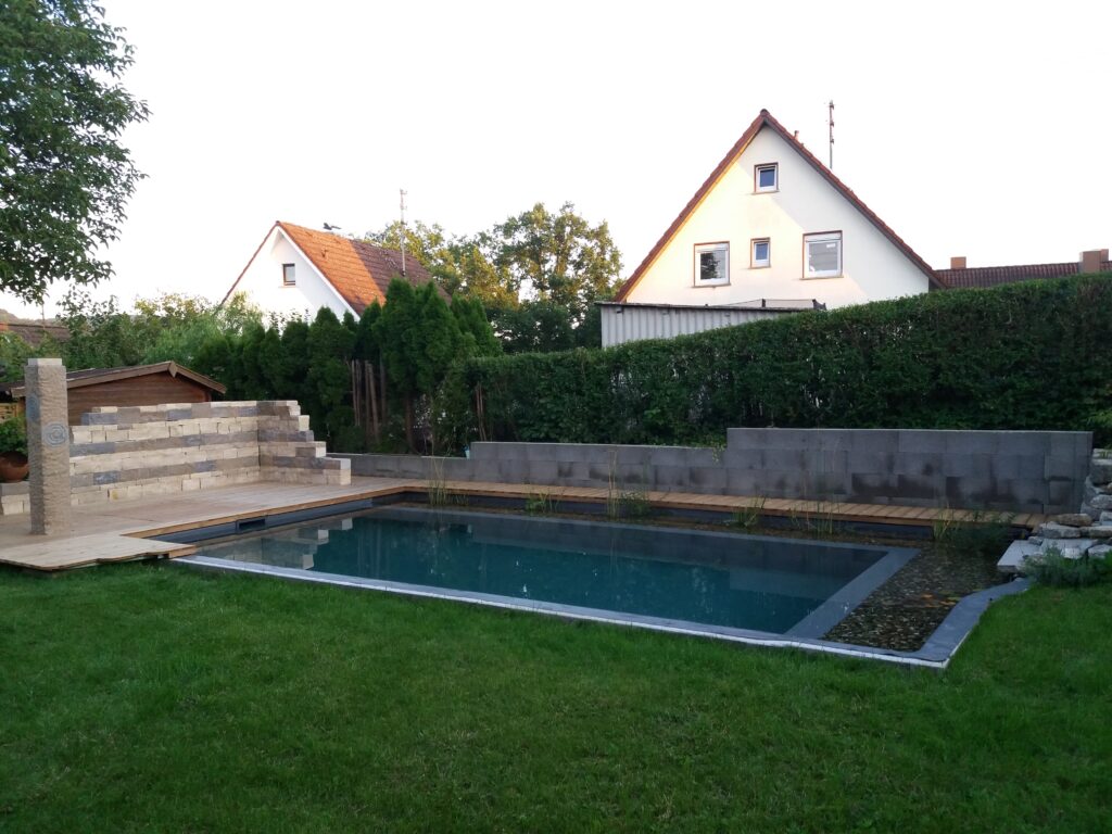 Naturpool ist von 2 Seiten mit Holz eingefasst. Auf der rechten Seite führt eine Treppe in die Filterzone als Einstieg in den Schwimmbereich.