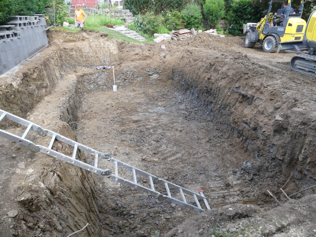 Aushub des Schwimmbereichs mit angelehnter Leiter, Steinmauer und Bagger im Hintergrund.