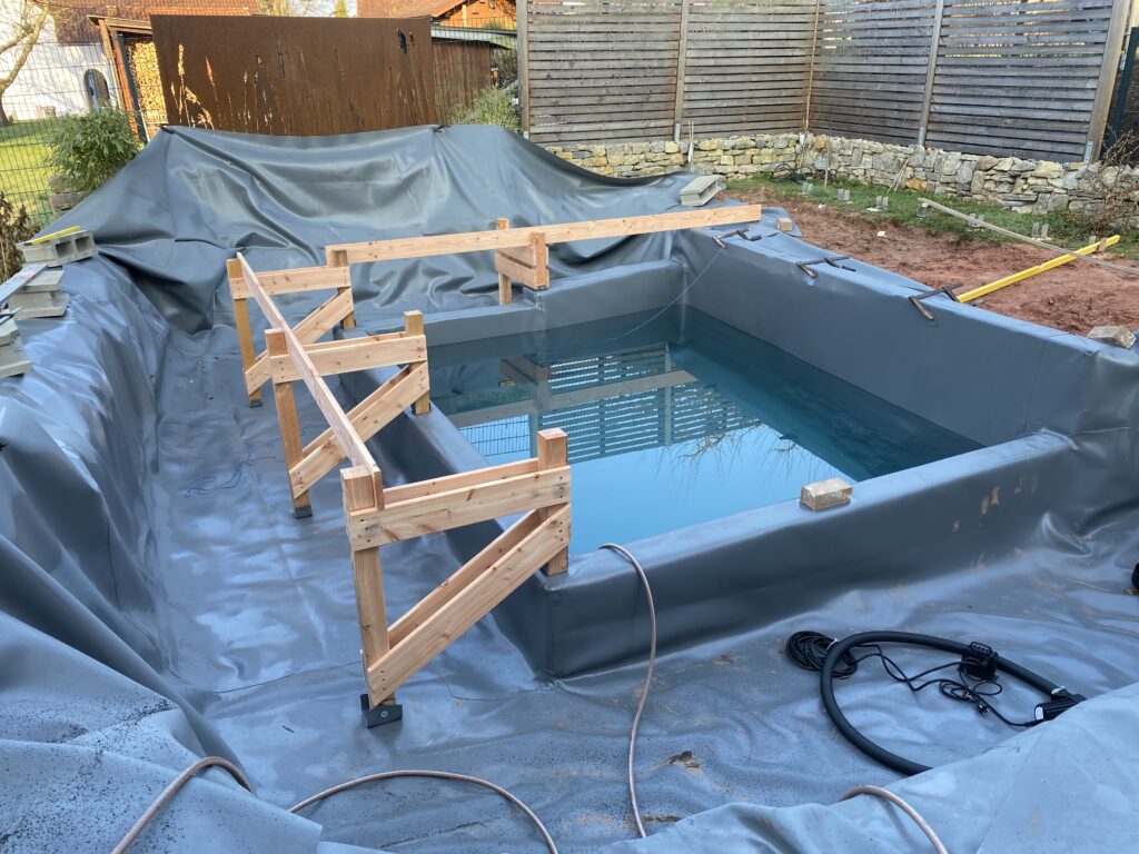 Der Schwimmbereich ist mit Wasser gefüllt und strahlt bläulich, während eine Holzkonstruktion in der Filterzone errichtet wird.