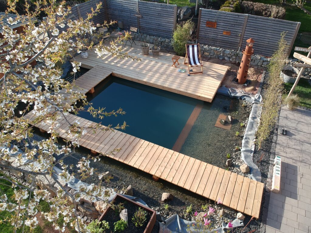 Eine Holzterrasse erstreckt sich über den Schwimmbereich, während der Holzweg um den Teich herumführt. Die Filterzone ist nun mit Wasser befüllt und bepflanzt, dank Mielke's Naturbadeteich.