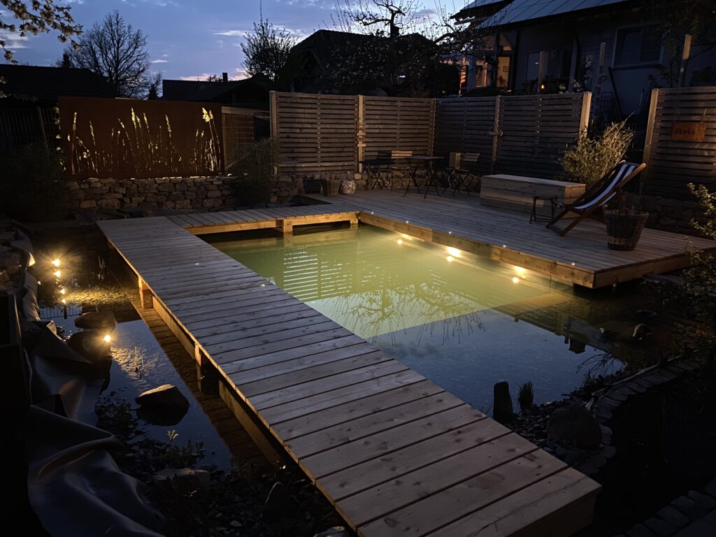 Ein beleuchteter Schwimmteich bei Abenddämmerung, ein Ergebnis der Arbeit von Mielke's Naturbadeteich beim Selbstbau des Schwimmteichs bei Fulda.