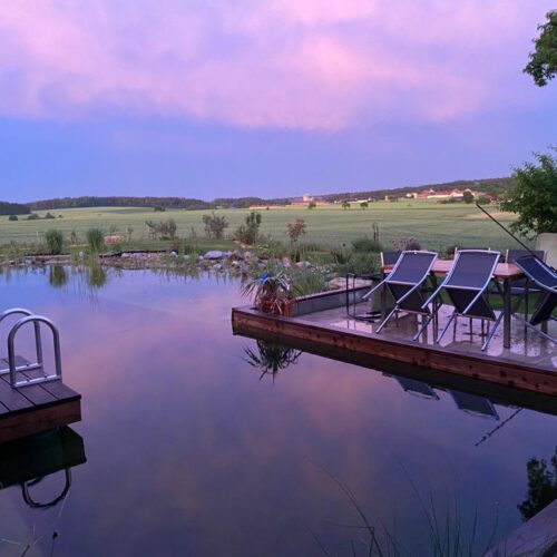 Ein romantischer Sonnenuntergang über dem Schwimmteich, mit den warmen Farben des Himmels, die sich im klaren Wasser widerspiegeln.
