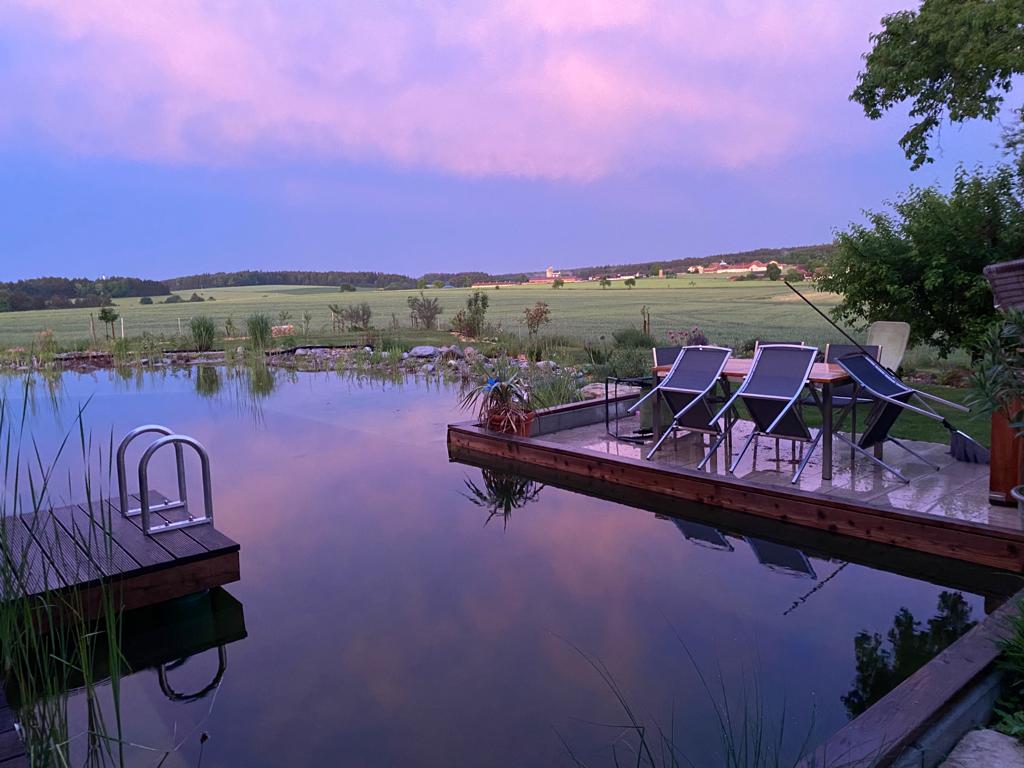 Ein romantischer Sonnenuntergang über dem Schwimmteich, mit den warmen Farben des Himmels, die sich im klaren Wasser widerspiegeln.