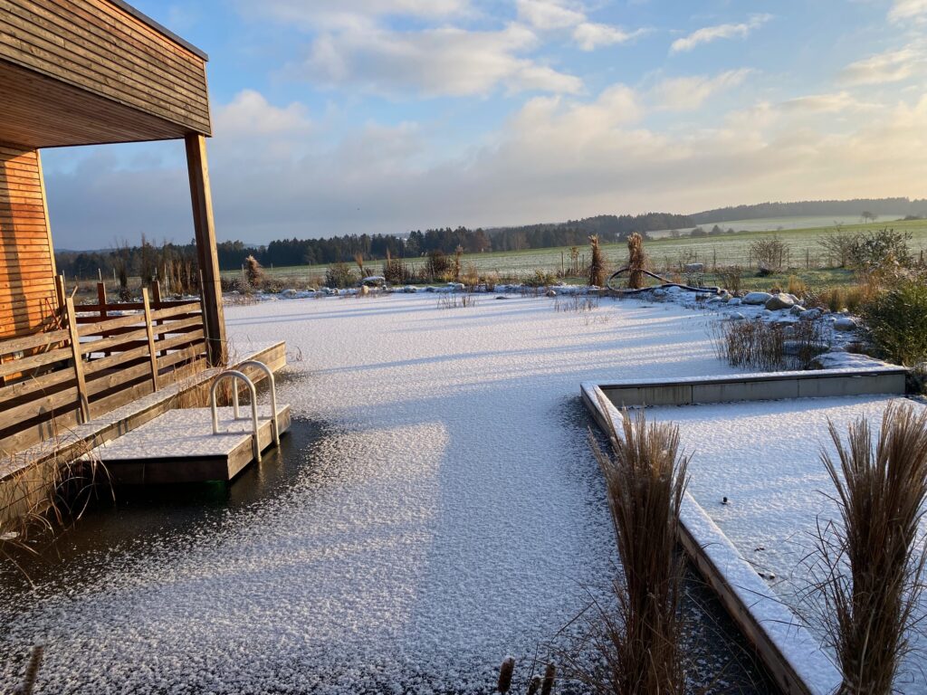 Verschneiter Schwimmteich im Sonnenlicht, Winterlicher Schwimmteich unter Sonnenstrahlen